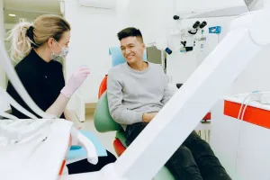 A dental professional, wearing a face mask and gloves, is interacting with a patient seated in a dental chair.